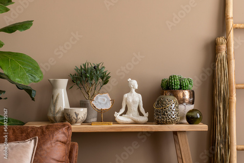 Ethnnic composition of living room interior with plants in flowerpots, brown sofa, wooden bench, ladder, round bowl, black ball, woman sculpture and personal accessories. Home decor. Template. photo