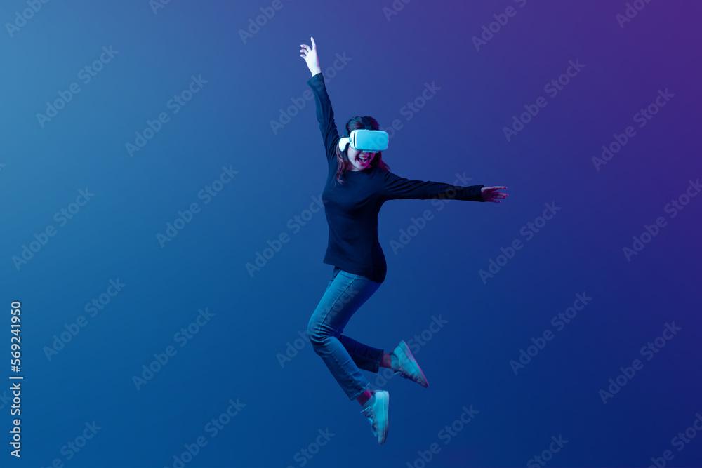 Isolated Young Asian woman wearing VR headset with experience playing video game and jumping levitating in the air on futuristic purple cyberpunk neon light background. Metaverse technology concept.