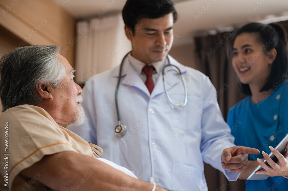 Smiling senior male patient lying and sitting on bed resting while nurse and doctor in lab coat and stethoscope showing digital tablet screen to man in a hospital room Smiling senior male patient lying and sitting on bed resting while nurse and doctor in lab coat and stethoscope showing digital tablet screen to man in a hospital room