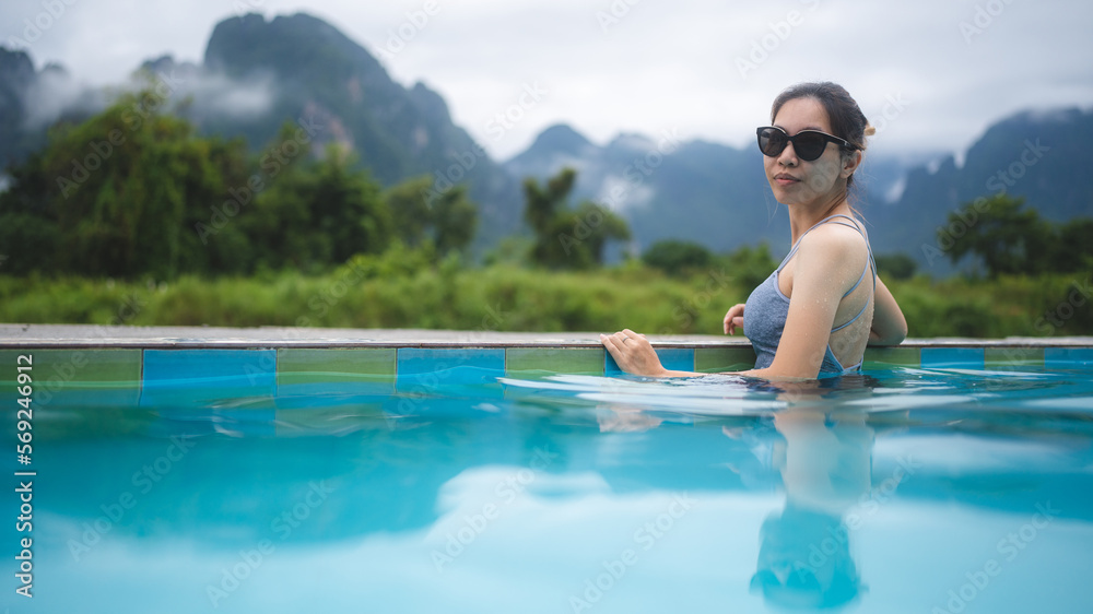 Rear view of young beautiful Asian woman in swimsuit relaxing while swimming in pool and leaning against edge and overlooking at the beautiful scenery while enjoying vacation