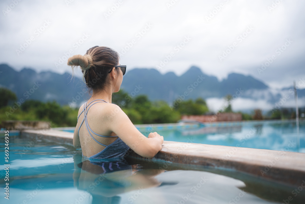 Rear view of young beautiful Asian woman in swimsuit relaxing while swimming in pool and leaning against edge and overlooking at the beautiful scenery while enjoying vacation