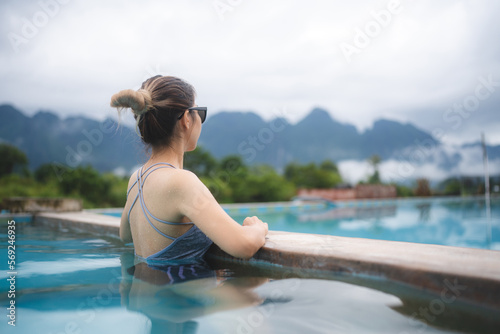 Rear view of young beautiful Asian woman in swimsuit relaxing while swimming in pool and leaning against edge and overlooking at the beautiful scenery while enjoying vacation © chokniti