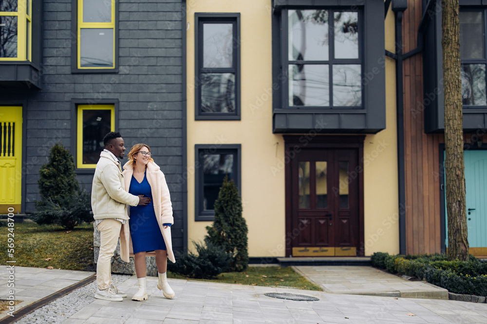 Young interracial couple stands and smiles on street against background of houses.