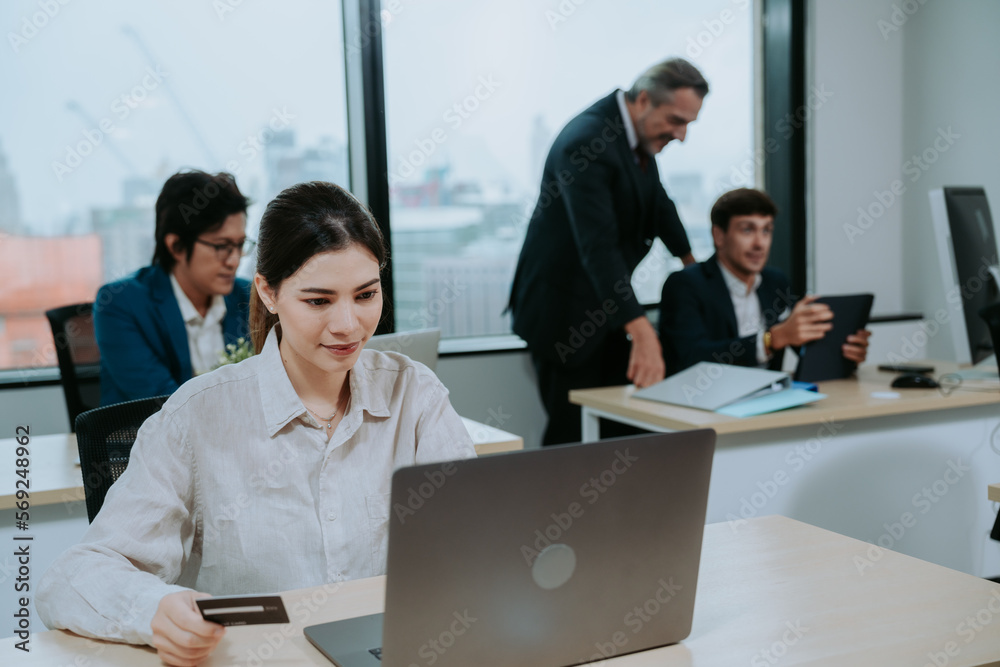 Focused and successful young female financer and marketing manager working and typing on laptop sitting in modern office with colleagues working in background Focused and successful young female financer and marketing manager working and typing on laptop sitting in modern office with colleagues working in background
