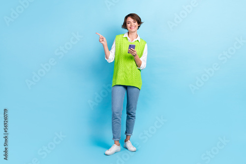 Full size photo of cute girl with brown hairdo wear green vest staring indicating empty space hold phone isolated on blue color background