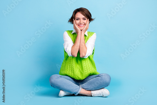 Full size photo of pleasant pretty cute girl with brown hairdo wear green vest jeans arms on cheekbones isolated on blue color background