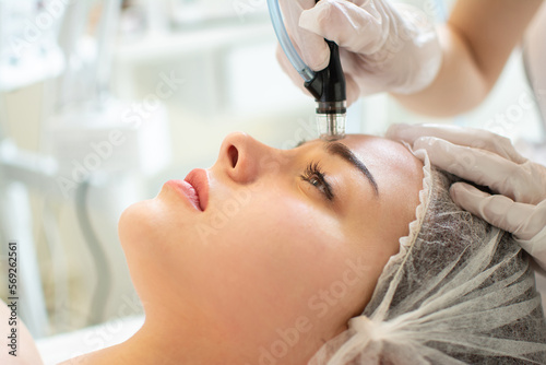 Close up of therapist's hands in protective gloves holding hydrafacial tool and performing anti aging progress to beautiful young woman forehead at beauty salon. photo