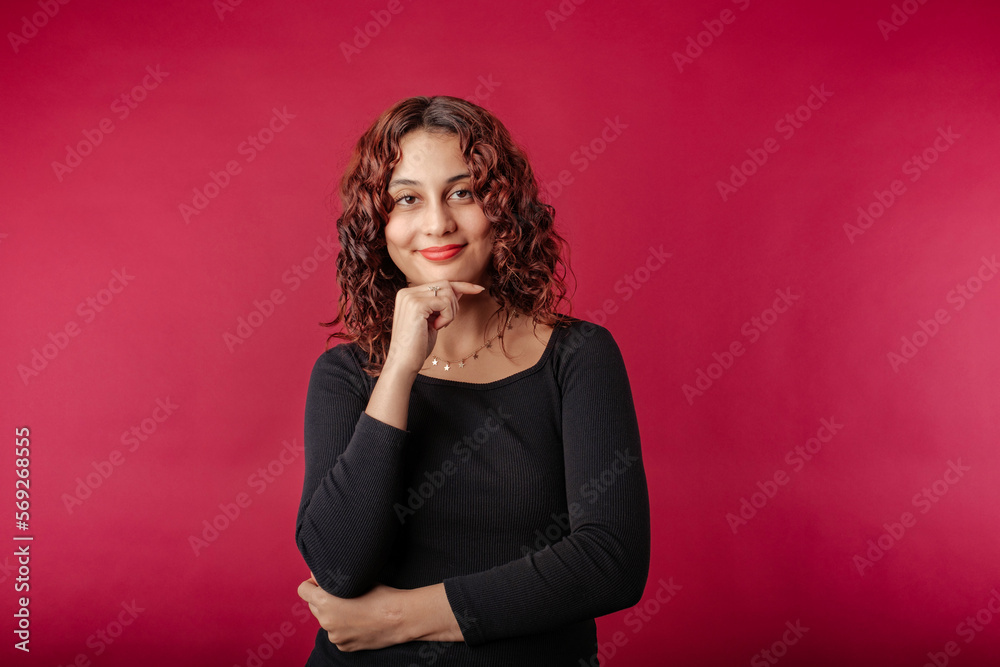 Fototapeta premium Beautiful redhead woman wearing black dress standing isolated over red background touches chin and looks suspiciously at the camera. Trying to find ideas.