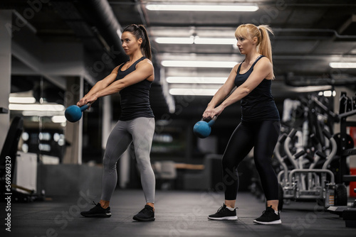 Two muscular sportswomen are swinging kettle bells in a gym.
