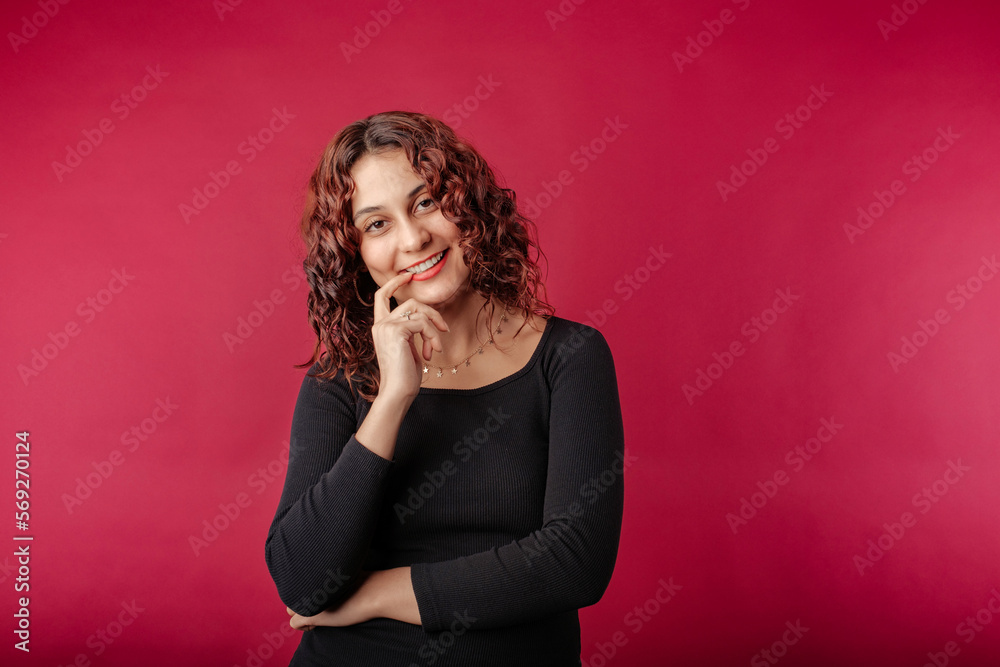 Fototapeta premium Young redhead girl smiling happy wearing black ribbed dress isolated over red background finger touch teeth ceramic wonder. Smiles at the camera with hope and joy.