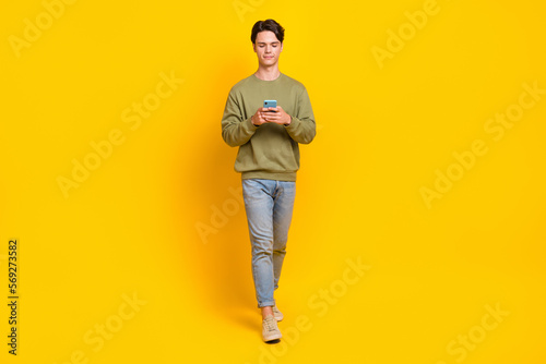 Full length portrait of nice young man hold use smart phone walking isolated on yellow color background