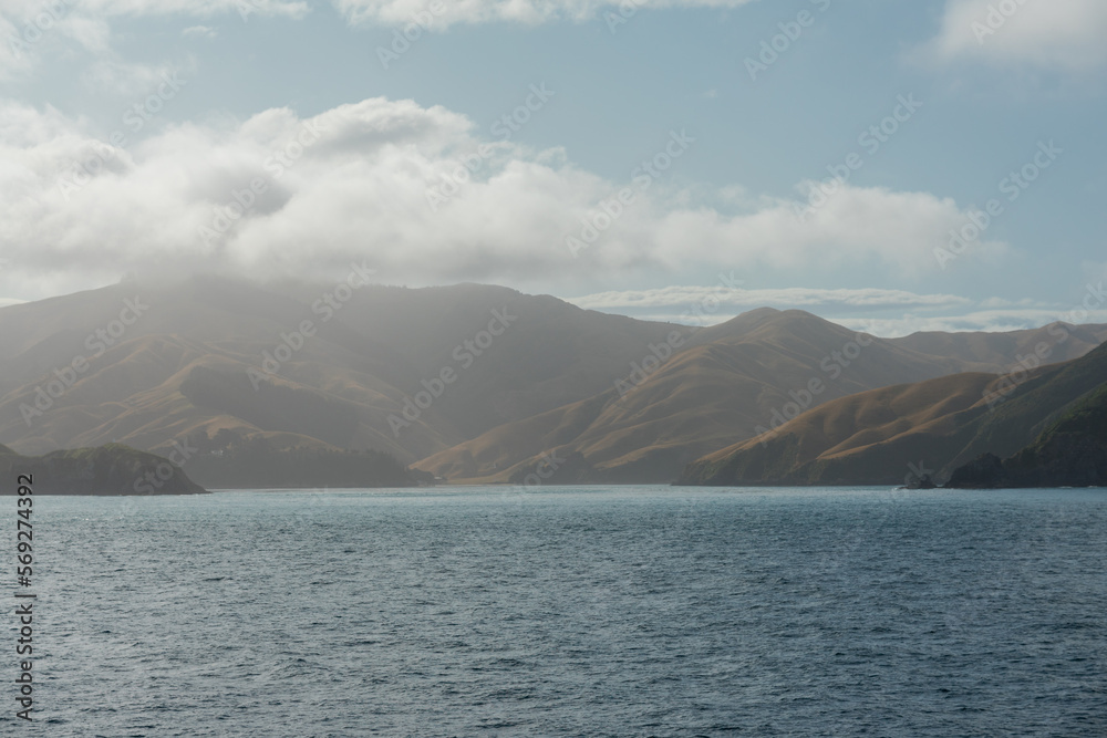 Marlborough Sounds in Neuseeland vom Meer aus in weichem Licht mit blauem Himmel und Sonne und Hügeln.