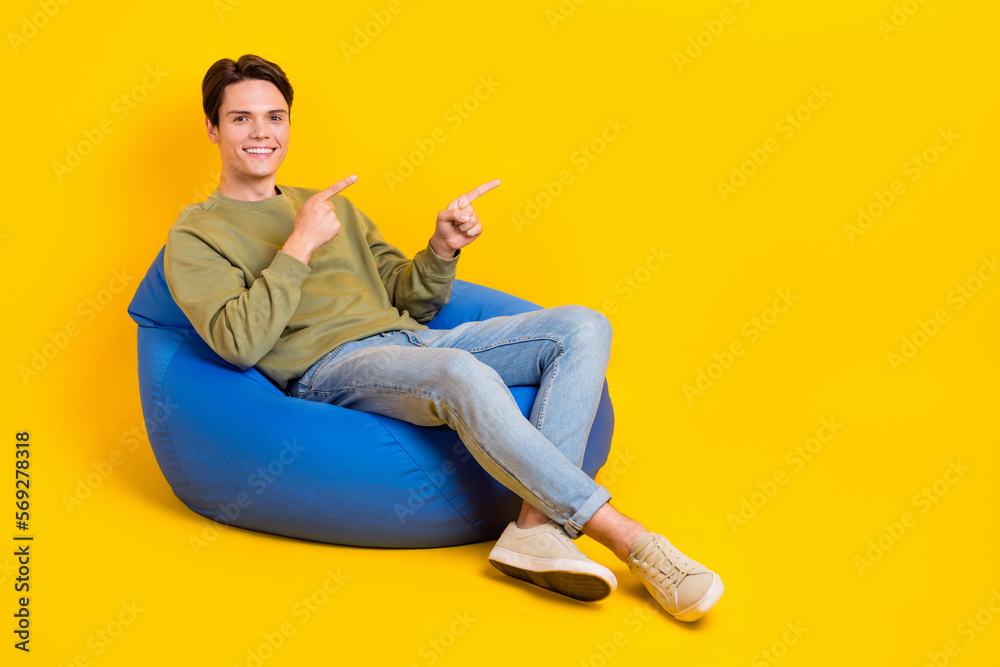 Full length portrait of positive cheerful person sit comfy bag direct fingers empty space isolated on yellow color background Full length portrait of positive cheerful person sit comfy bag direct fingers empty space isolated on yellow color background