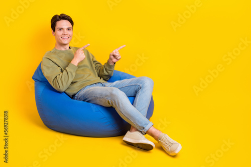 Full length portrait of positive cheerful person sit comfy bag direct fingers empty space isolated on yellow color background