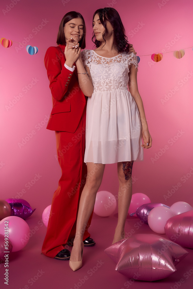 Valentine's day concept. Studio portrait of two young lesbian girls holding hands. One girl is in a white dress, the second woman is in a red suit. They stand on a pink background with balloons. Valentine's day concept. Studio portrait of two young lesbian girls holding hands. One girl is in a white dress, the second woman is in a red suit. They stand on a pink background with balloons.
