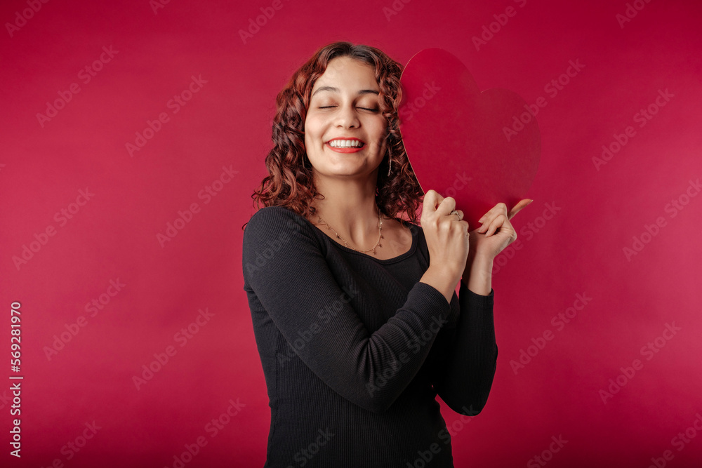 Beautiful redhead woman wearing dress standing isolated over red background smiling with love in a warm, friendly pose with a large cardboard heart shape. Cuddle big paper card heart shape figure. Beautiful redhead woman wearing dress standing isolated over red background smiling with love in a warm, friendly pose with a large cardboard heart shape. Cuddle big paper card heart shape figure.