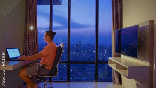 Handsome man in room luxury hotel works at laptop. Nice guy lives or freelancing works in hotel. Man in an apartment against background of night city. Man sits on an office chair against large window