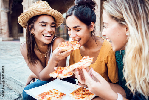 Three cheerful multiracial young women eating italian pizza at city street. Young female friends enjoying summer holidays. Vacation lifestyle concept