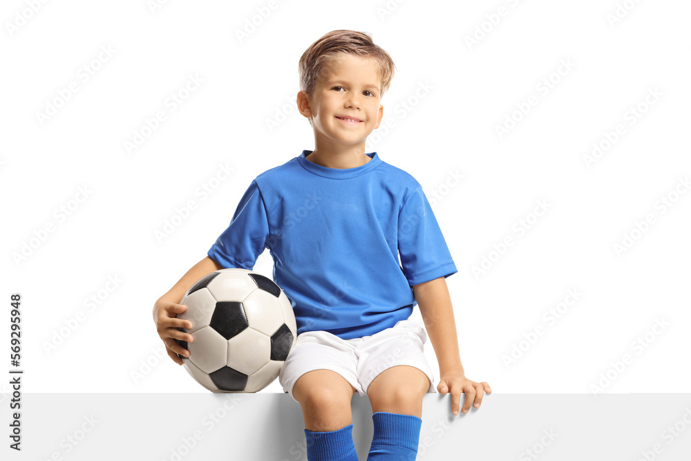 Boy in a blue soccer jersey sitting on a blank panel and holding a ball