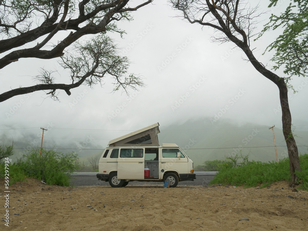 A pop-up campervan is parked at a beach in Hawaii with low cloud hanging in the hills in the background 