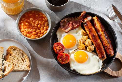 Full English breakfast with fried eggs, sausages, beans, bacon, grilled tomatoes, mushrooms, toasts and cup of coffee on gray background.