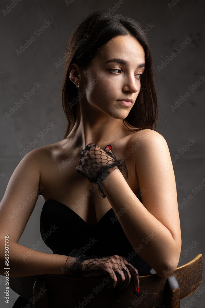 Classic dark studio portrait of a young brunette woman in black clothes who is sitting on a chair.
