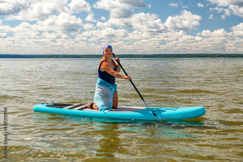 A Jewish woman in a headscarf and a pareo on her knees on a SUP board with an oar swims in a lake on a sunny day.