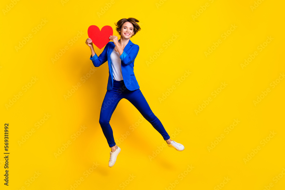 Full length photo of charming cute lady dressed blue blazer jumping high holding heart empty space isolated yellow color background