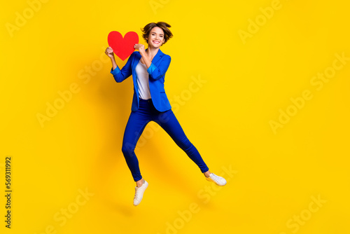 Full length photo of charming cute lady dressed blue blazer jumping high holding heart empty space isolated yellow color background