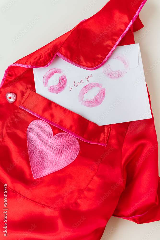 The inscription I love you on a white envelope with a print of lips made with red lipstick on a background of small red hearts and a red shirt. Valentine concept, vertical photo.