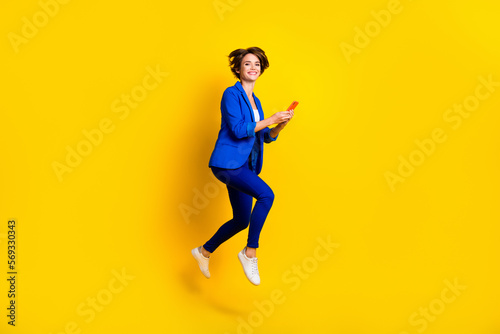 Full length photo of sweet cute lady dressed blue blazer jumping high chatting modern gadget isolated yellow color background