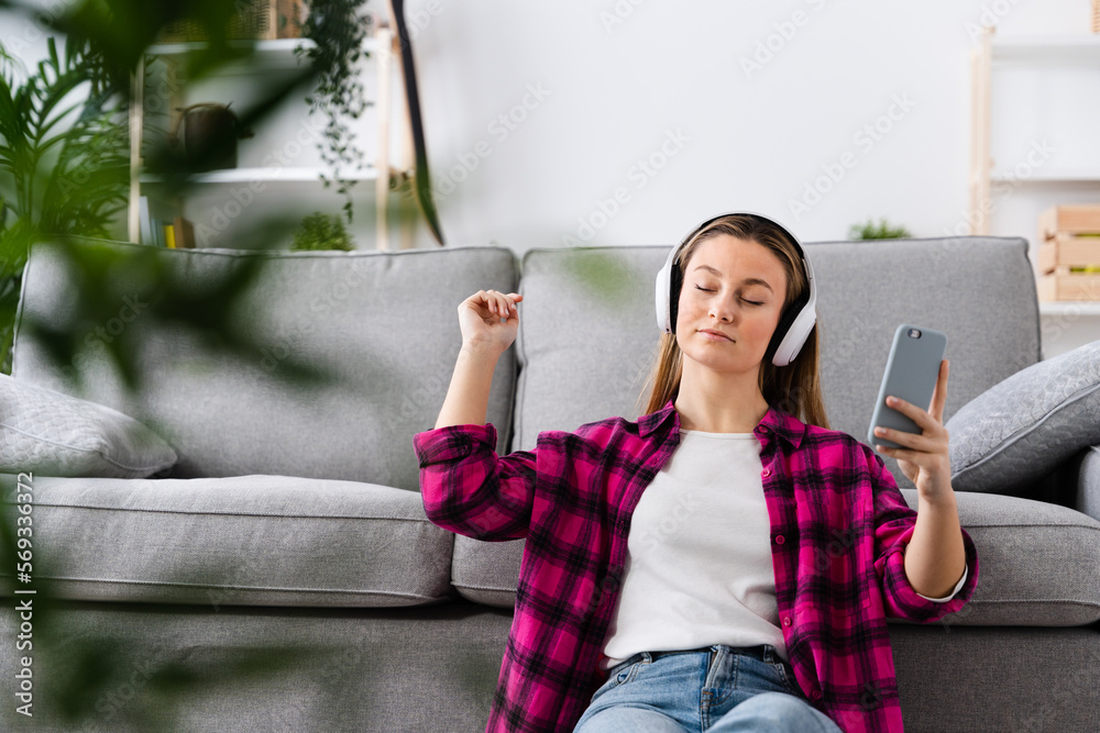 Young pretty woman listening to music in living room and holding phone