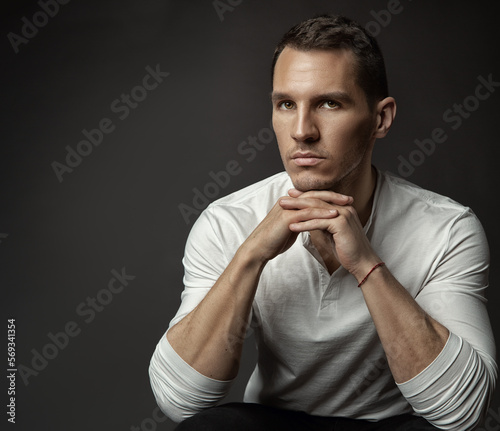 Handsome Young Man. Elegant Fashion Men Face Portrait looking at camera in White Shirt over dark Gray Background. Serious beautiful Unsaved Male Model with Crossed hands fingers Under Chin © inarik