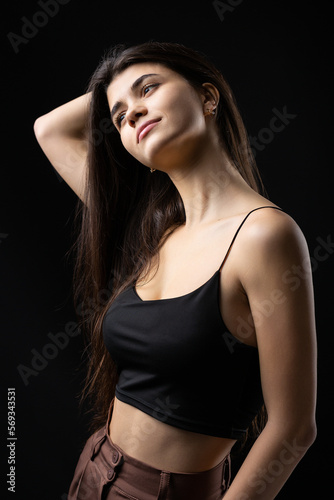 Classic studio portrait of a young brunette dressed in a black top, who is sitting on a chair against a black background.