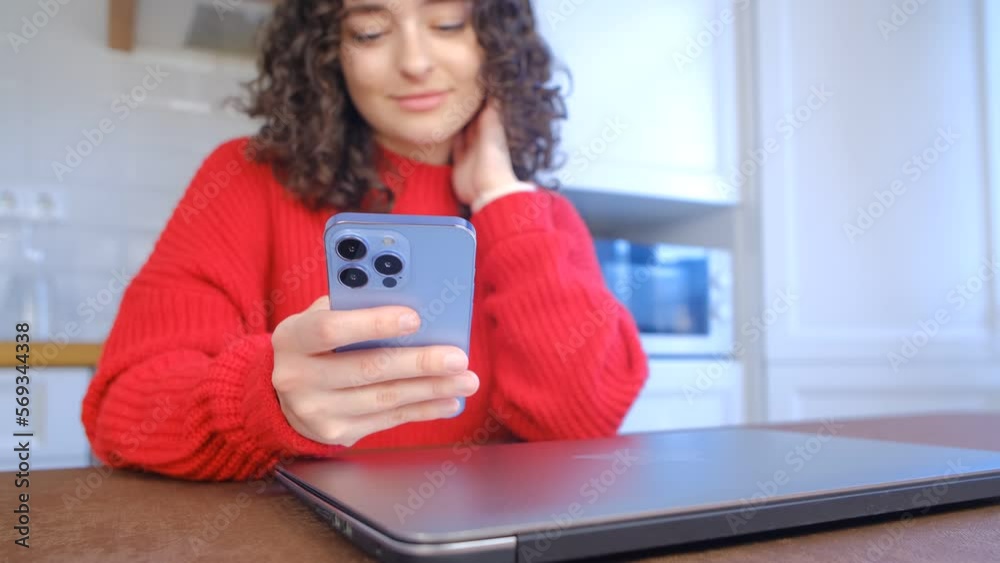 Beautiful young girl with curly hair browsing mobile app on a smartphone. Cheerful Ukrainian female person using modern mobile phone at home Beautiful young girl with curly hair browsing mobile app on a smartphone. Cheerful Ukrainian female person using modern mobile phone at home