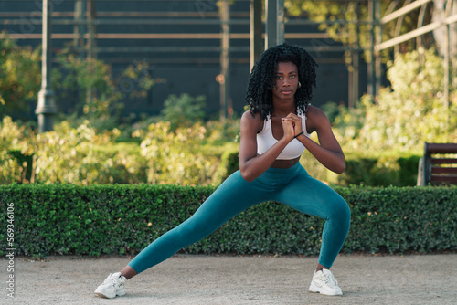 mujer de color negra con ropa deportiva, preparándose para hacer ejercicios, sentadillas, estirando, flexiones, corriendo en un parque con luz natural. 