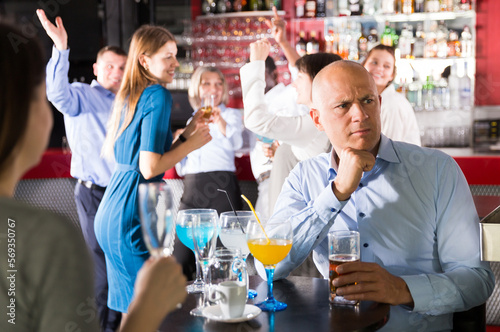 Troubled man sitting alone at office party in nightclub on background with cheerful workmates