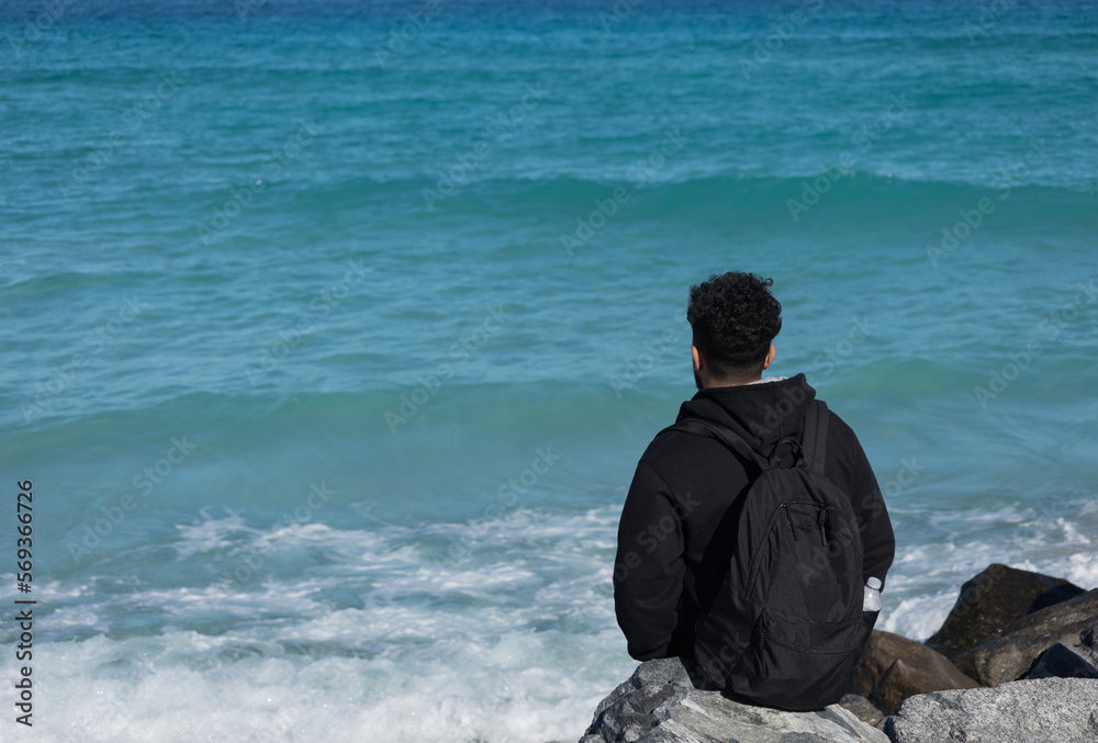 A young guy with a backpack sits on the rocks on the ocean. Loneliness of men. Tourist. Soft focus A young guy with a backpack sits on the rocks on the ocean. Loneliness of men. Tourist. Soft focus