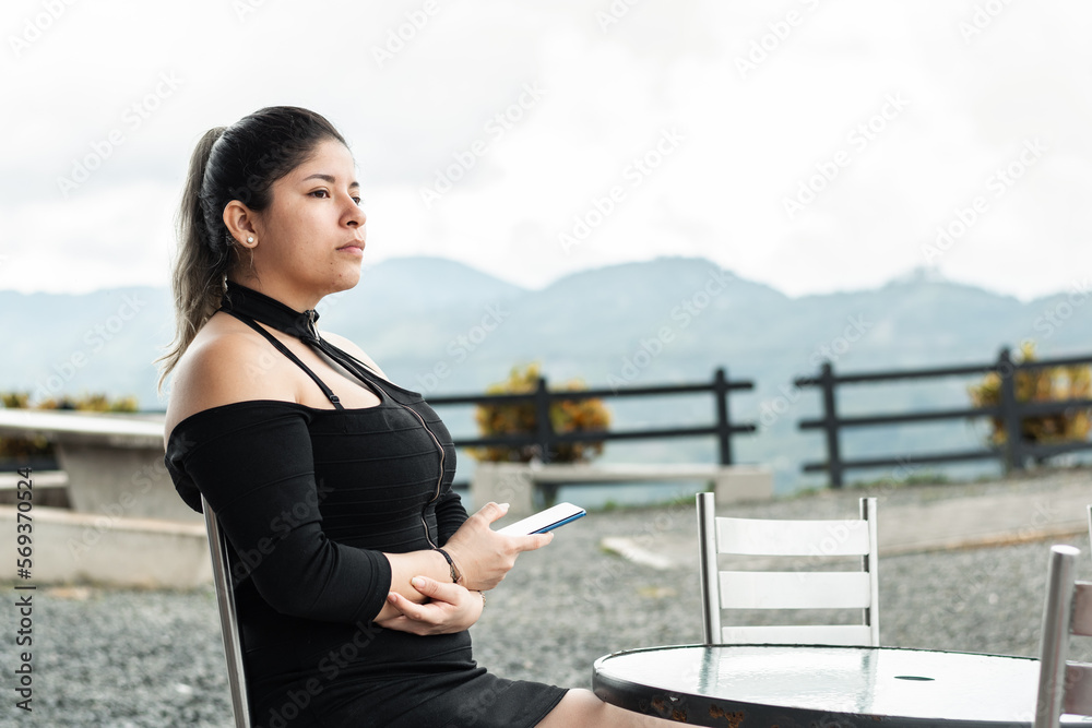 young latina woman, cell phone in hand, sitting waiting in an outdoor restaurant