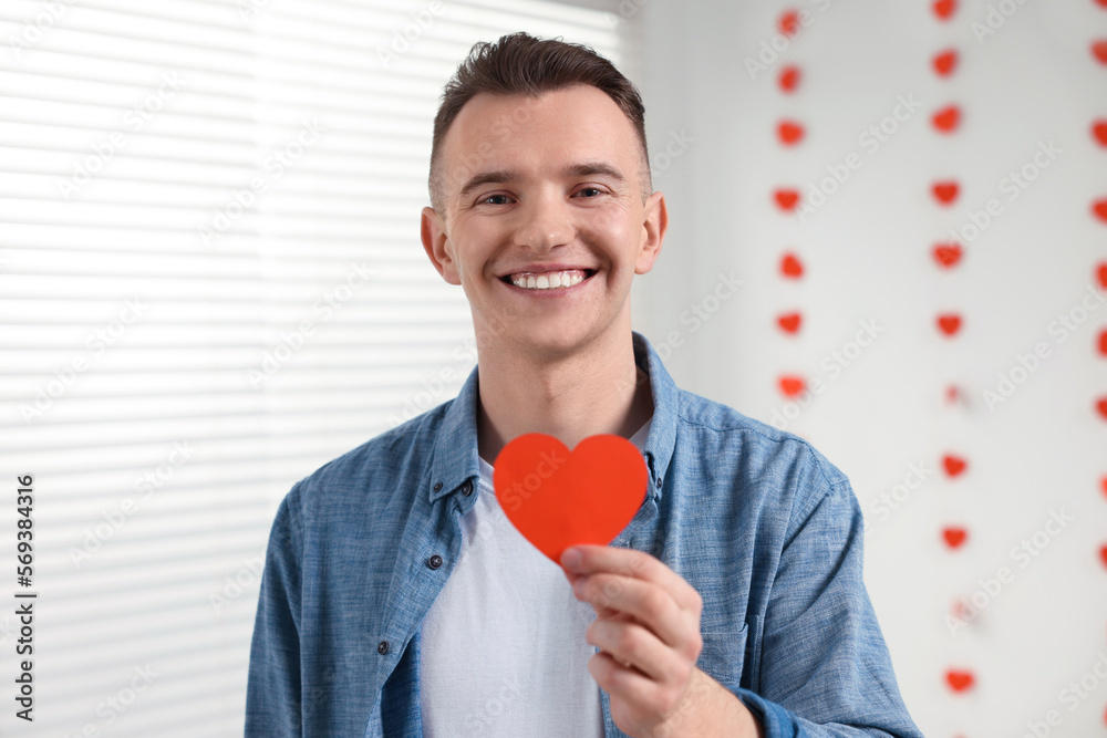 Handsome young man with decorative heart indoors, view from camera. Valentine's day celebration in long distance relationship Handsome young man with decorative heart indoors, view from camera. Valentine's day celebration in long distance relationship