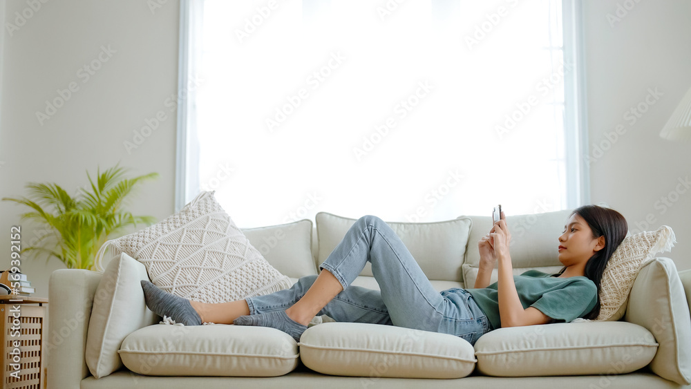 Happy young asian woman relaxing at home. Female is lying down on sofa and using mobile smartphone Happy young asian woman relaxing at home. Female is lying down on sofa and using mobile smartphone