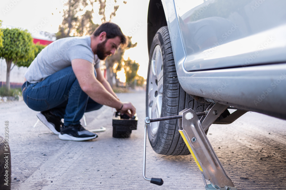 young man loosening a nut of his car tire, changing a flat tire on the road, car jack being used young man loosening a nut of his car tire, changing a flat tire on the road, car jack being used