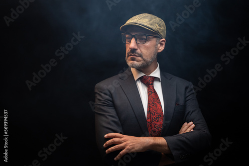 Portrait of detective in elegant black suit and red tie wearing eye glasses and hat standing with crossed arms. isolated on black background.