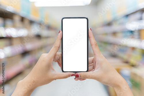 Close up of two female hands holding a mobile phone on a vertical blank white screen. Blurred supermarket background. Mockup.