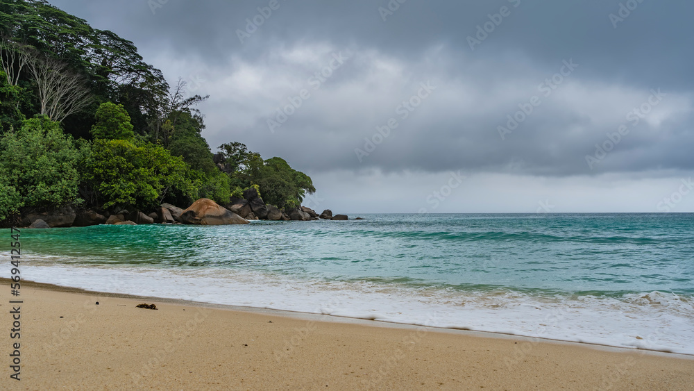 Fototapeta premium Rainy foggy day on a tropical island. The waves of the turquoise ocean are foaming on the sand of the beach. A hill overgrown with green vegetation, against a cloudy sky. Boulders in the water.