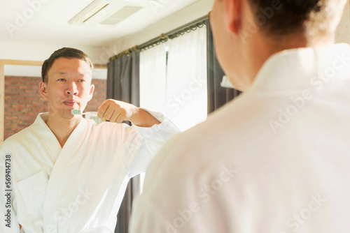 Asian man in white bathrobe brushing teeth standing in front of mirror  oral care concept.