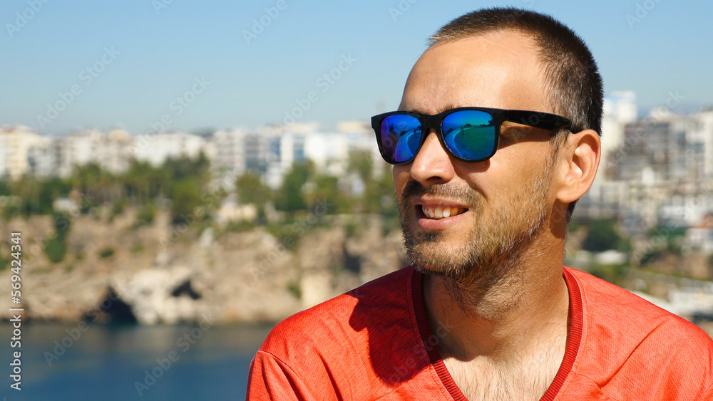 A handsome man with sunglasses smiling against the background of the sea and the city. Caucasian man in red jersey against a beautiful seascape with mountains.