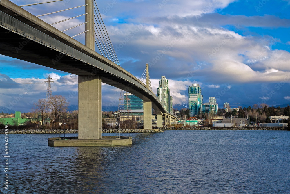 Fototapeta premium Sky-train bridge linking Richmond and Vancouver City over the Fraser River, city on the opposite bank, stormy sky with clouds on a background