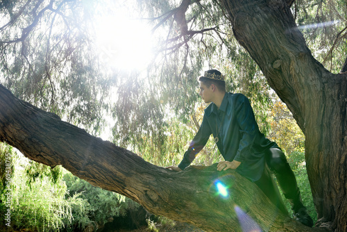 portrait of handsome masked man wearing fantasy medieval prince costume with golden crown and romantic silk shirt.  climbing a tree branch in a forest location with backlit silhouette lighting.
