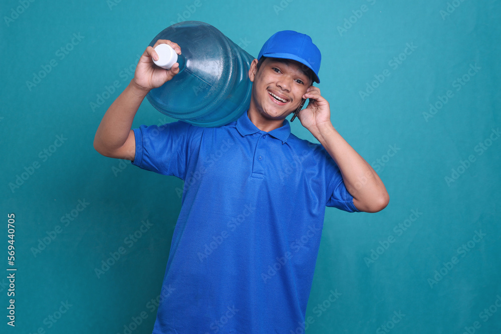 Asian delivery guy in blue uniform work as dealer courier, holding water bottle while doing a phone call Asian delivery guy in blue uniform work as dealer courier, holding water bottle while doing a phone call